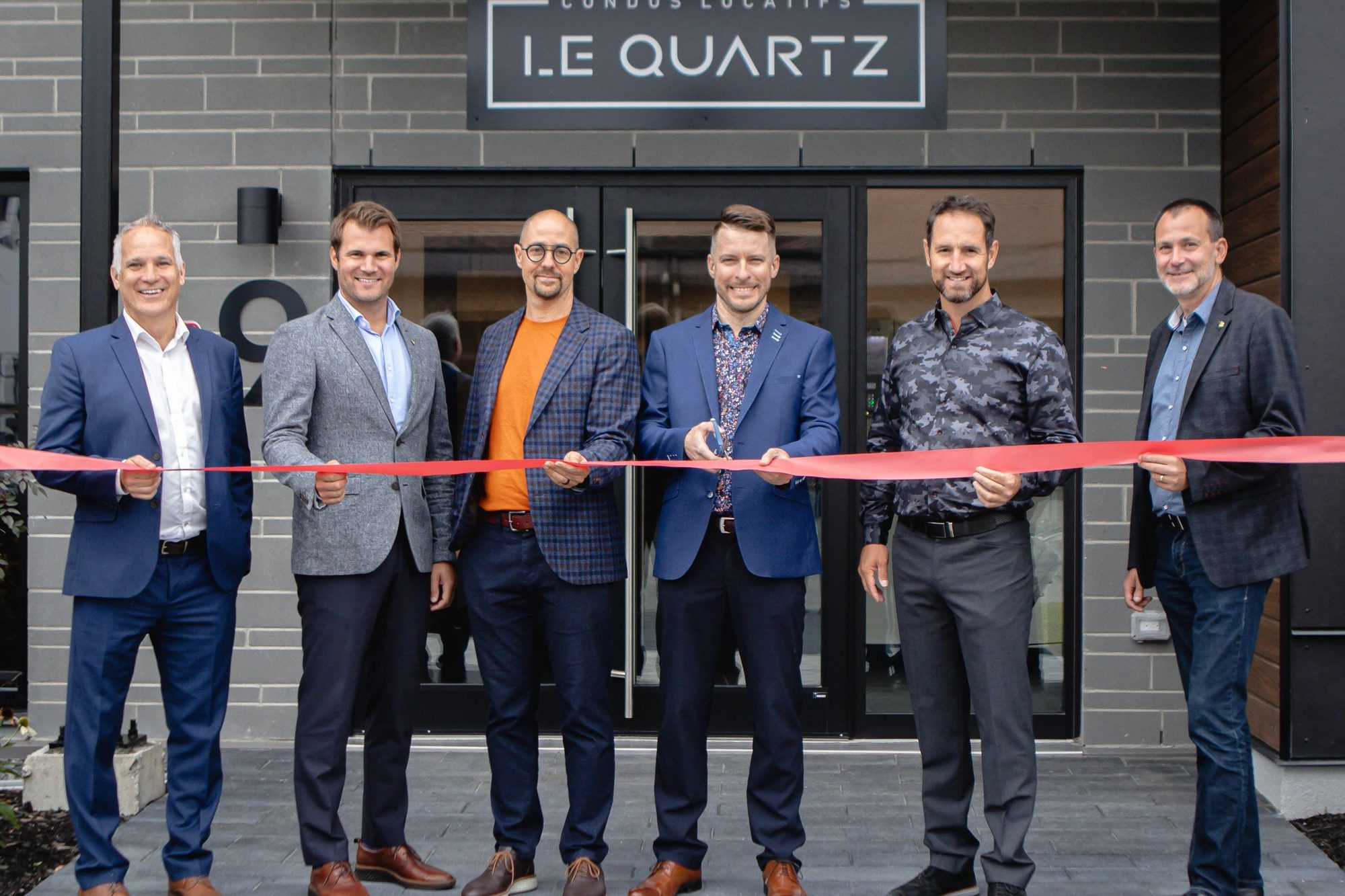 Six hommes en veston coupent un ruban rouge devant l'entrée du Quartz, sous l'enseigne Condos locatifs Le Quartz