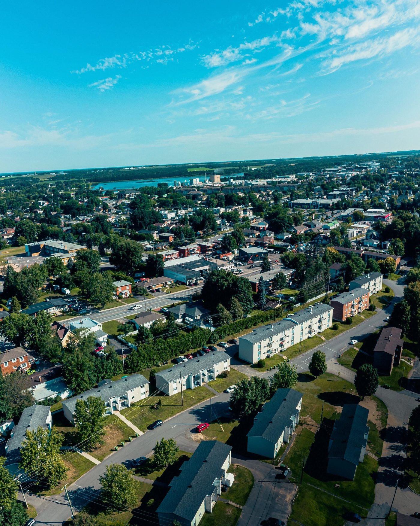 Vue aérienne d'un quartier résidentiel de Victoriaville en été : des immeubles de logements de deux étages entre les arbres, la ville et la rivière au fond