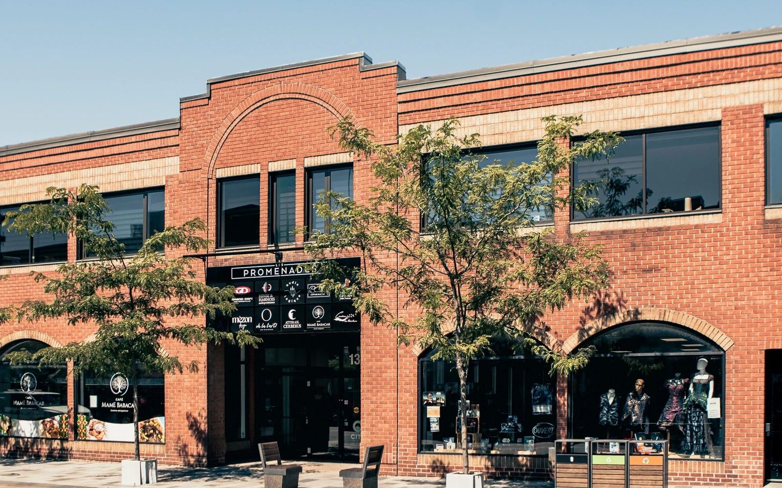 L'entrée des Promenades au centre-ville : l'enseigne noire sous l'arche de brique, la liste des commerces, un arbre devant